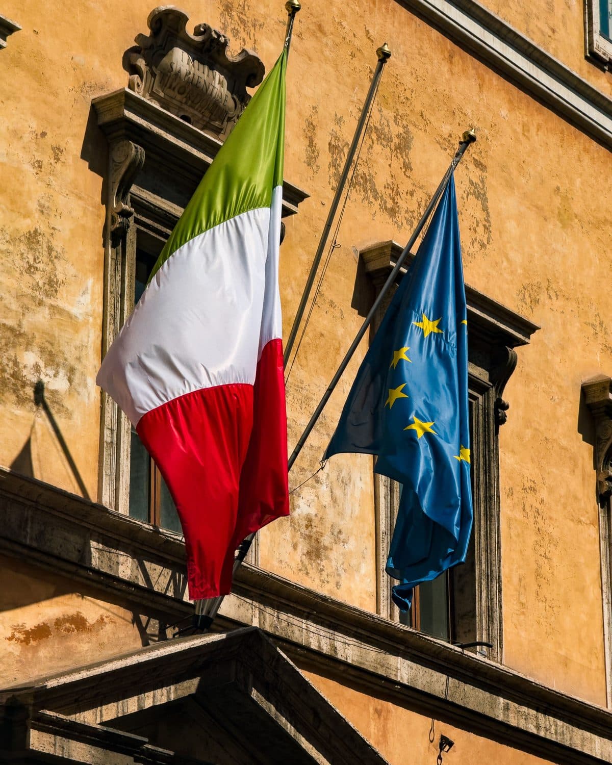 The Italian and European Union flags on the facade of an ochre-rendered building.
