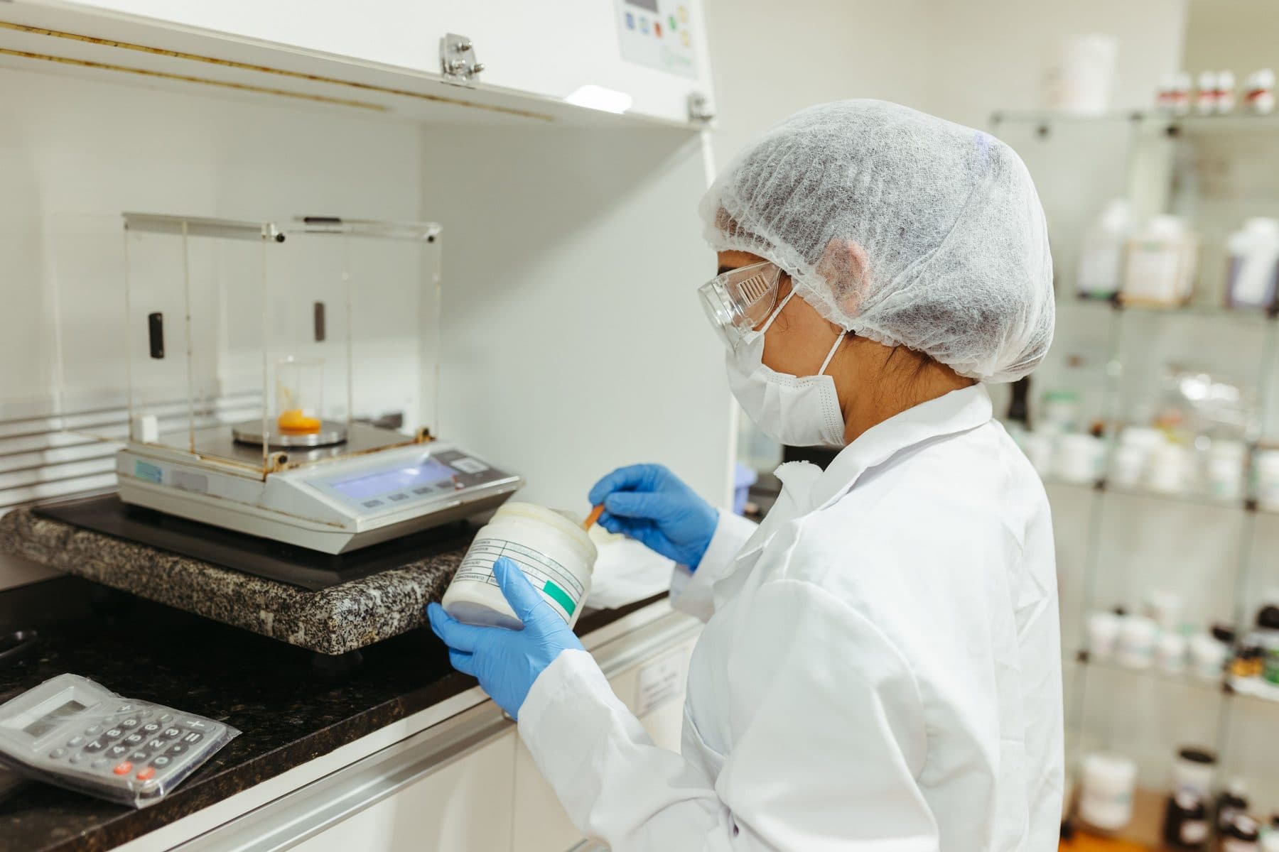 A technician in a hairnet, mask and gloves weighing a powdered ingredient on a laboratory balance.