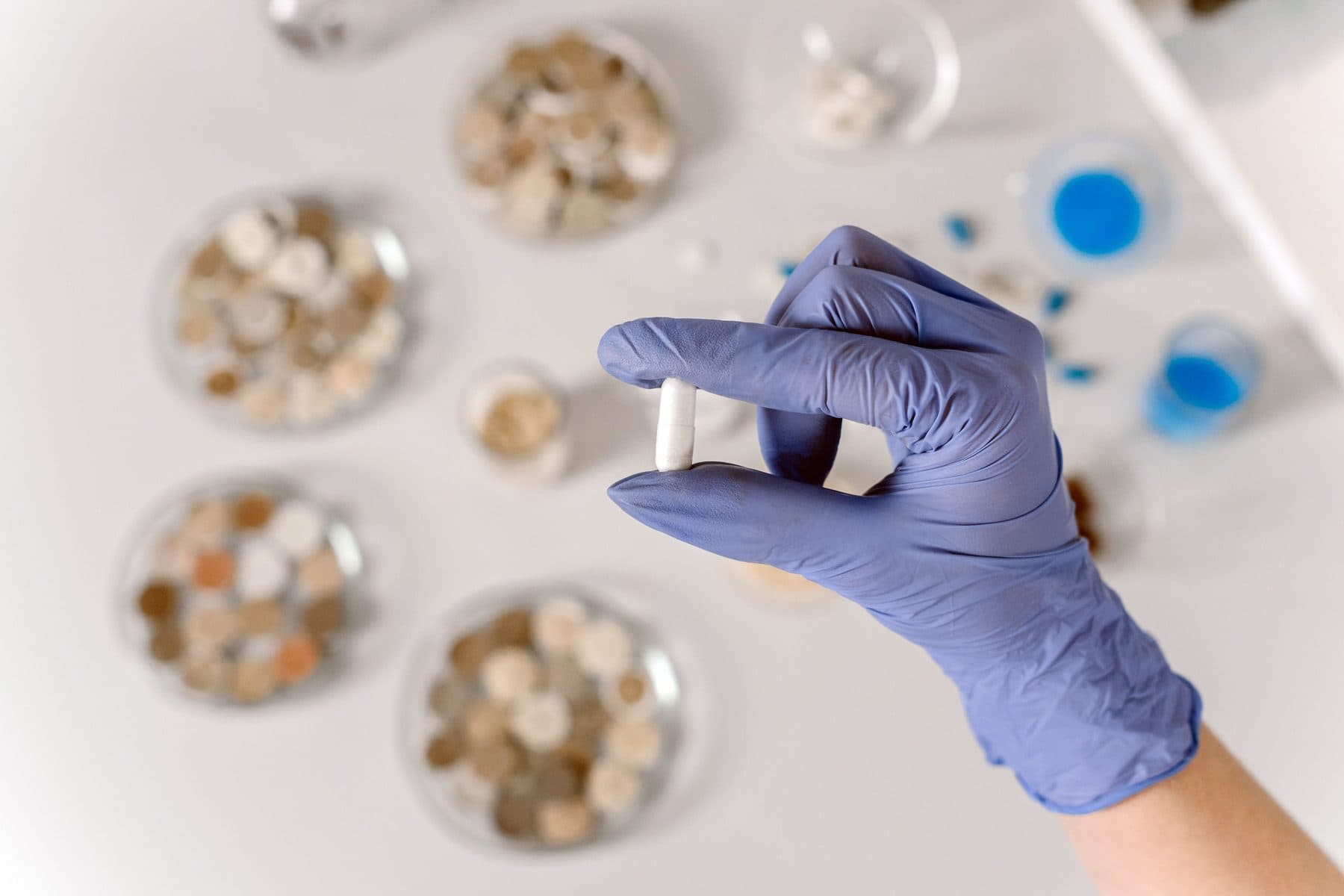 A gloved hand holding a single white capsule above dishes of raw ingredients.